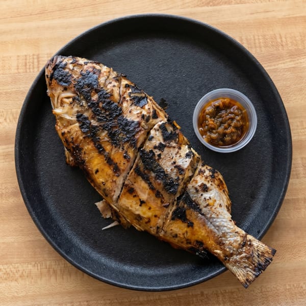 Grilled whole fish with dark char marks, served on a black plate with a side of sauce.