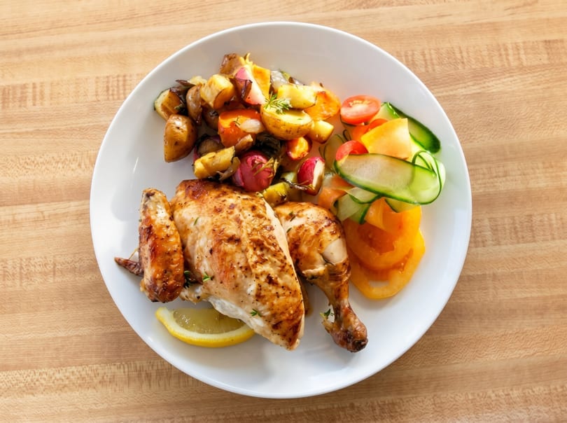 Roasted chicken with potatoes, radishes, and a side salad of tomatoes and cucumber on a white plate.