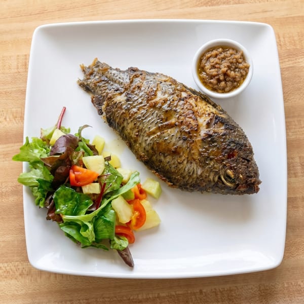 Whole grilled fish with a side salad of greens, tomatoes, and potatoes, and a small bowl of sauce.
