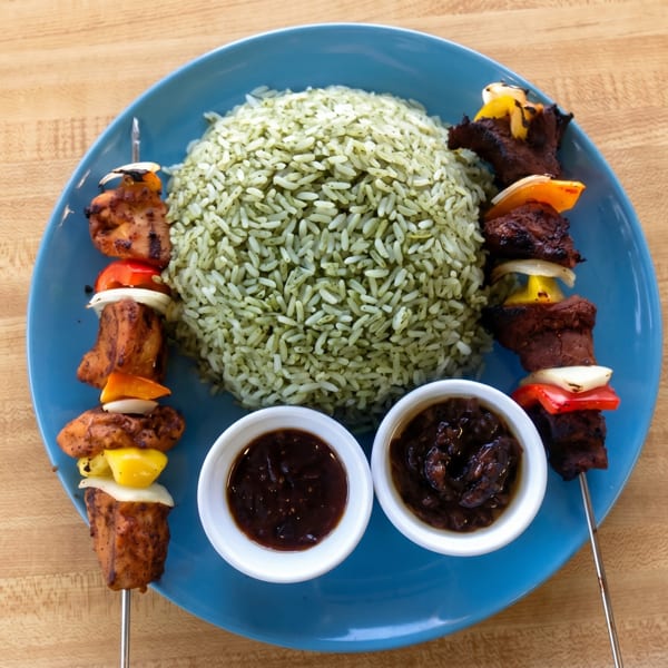 Two skewers of grilled meat and vegetables, green rice, and two dipping sauces on a blue plate.
