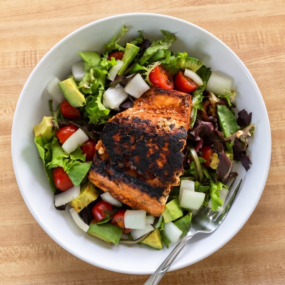 Grilled salmon salad with mixed greens, avocado, cherry tomatoes, and onions in a white bowl.