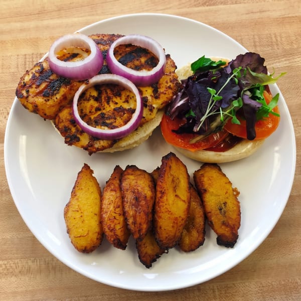 Grilled chicken sandwich with red onion, tomato, mixed greens, and a side of fried plantains.