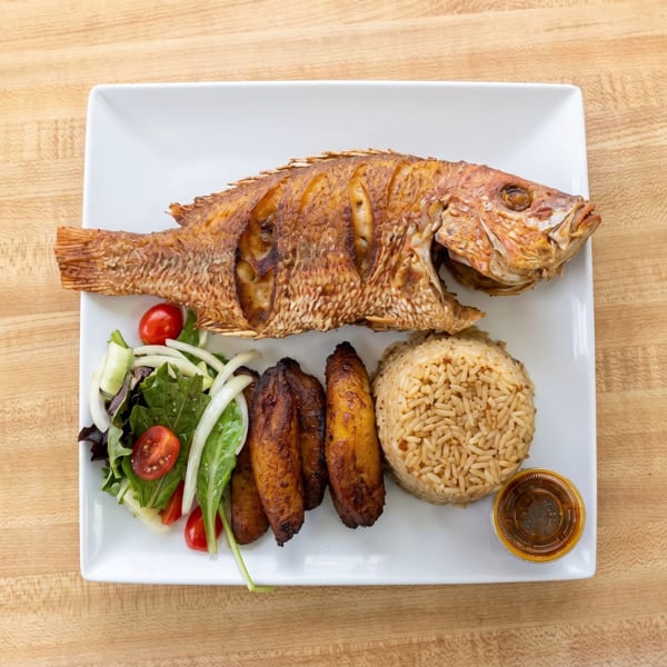 A whole fried fish with rice, plantains, and a side salad on a white rectangular plate.